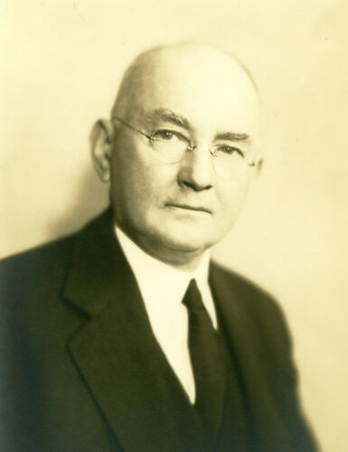 Photograph: A sepia-toned portrait of a man wearing a white shirt, black suit and tie, and glasses.