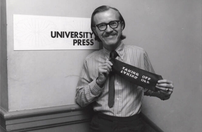 Photo: A smiling man in 1970s-era attire holds up his necktie, which has been stenciled with the words 'Taking Off.' He stands in front of a sign that reads 'University Press.'