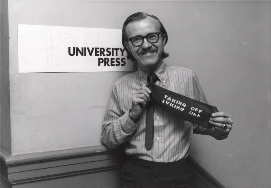 Photo: A smiling man in 1970s-era attire holds up his necktie, which has been stenciled with the words 'Taking Off.' He stands in front of a sign that reads 'University Press.'